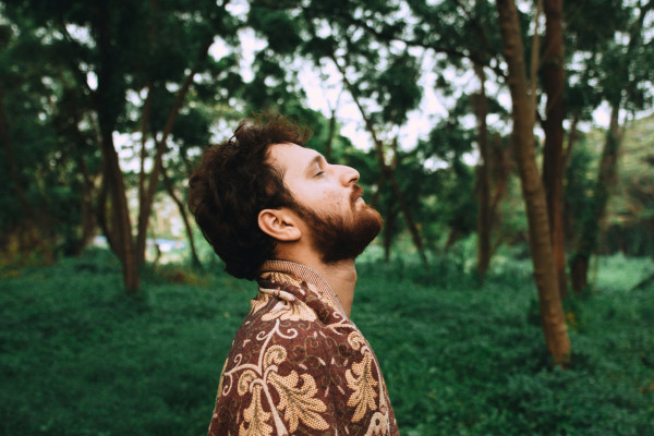 A man stands calmly near trees in a forest, taking a quiet moment to breathe.