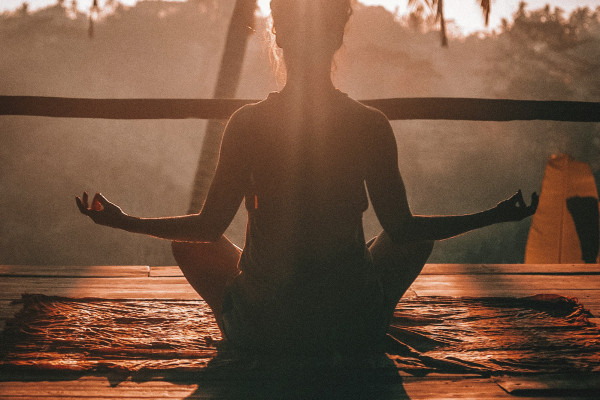 A woman sitting cross-legged on a wooden floor with her eyes closed, hands resting gently in a meditative pose.