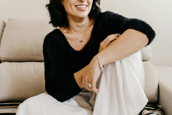 A woman sitting thoughtfully on a white couch, wearing a black long-sleeve shirt.