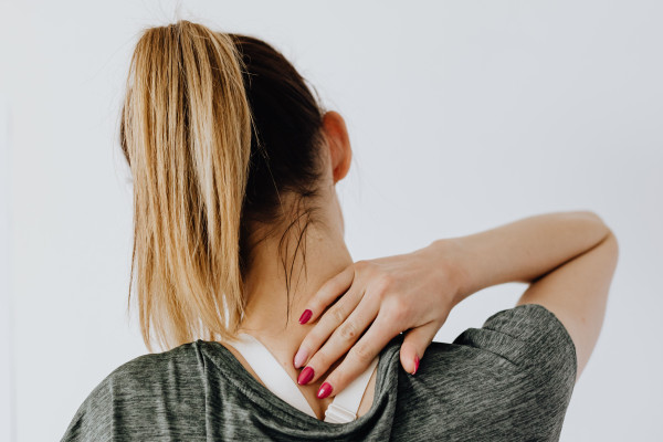 Woman from behind holding the back of her neck with one hand, showing signs of neck discomfort while sitting indoors.