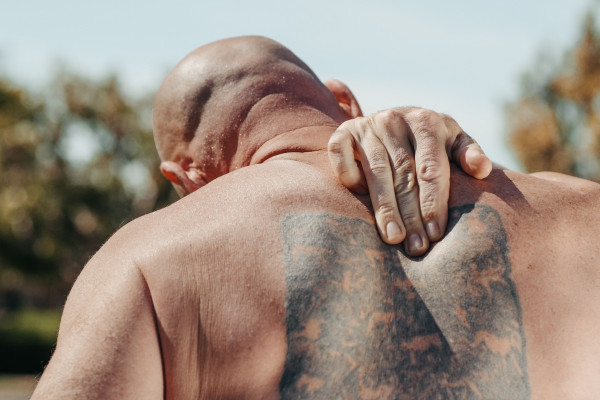 Bald man with a large back tattoo holding the back of his neck outdoors, showing signs of discomfort
