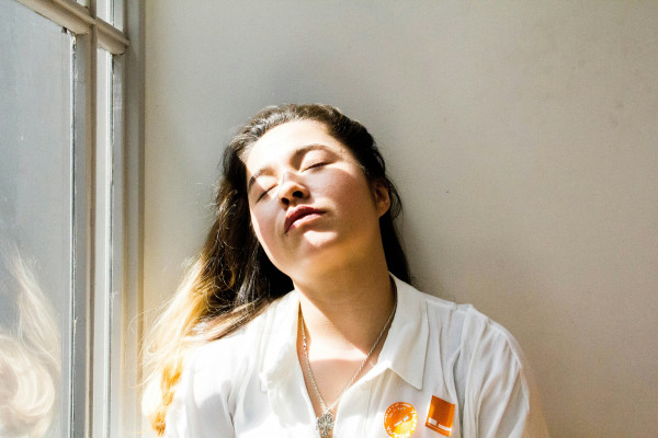 A woman standing quietly near a window, looking tired and reflective in soft daylight.
