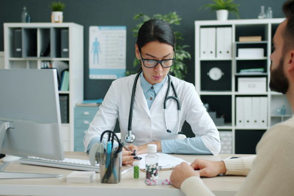 Clinician sitting at a desk, talking with a patient and taking notes during a health consultation